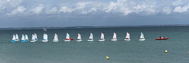 Photo von sechzehn kleinen Segelbooten in einer Reihe auf dem Meer. Vorne weg fährt ein rotes Motorschlauchboot.