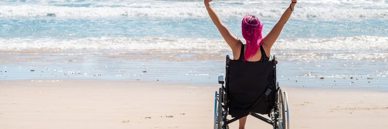 Frau im Rollstuhl, die ihre Arme mit zwei Fäusten nach oben streckt, am Strand und blickt zum Meer.